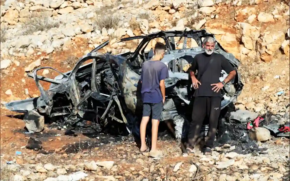 People stand next to a car destroyed by an Israeli air strike
